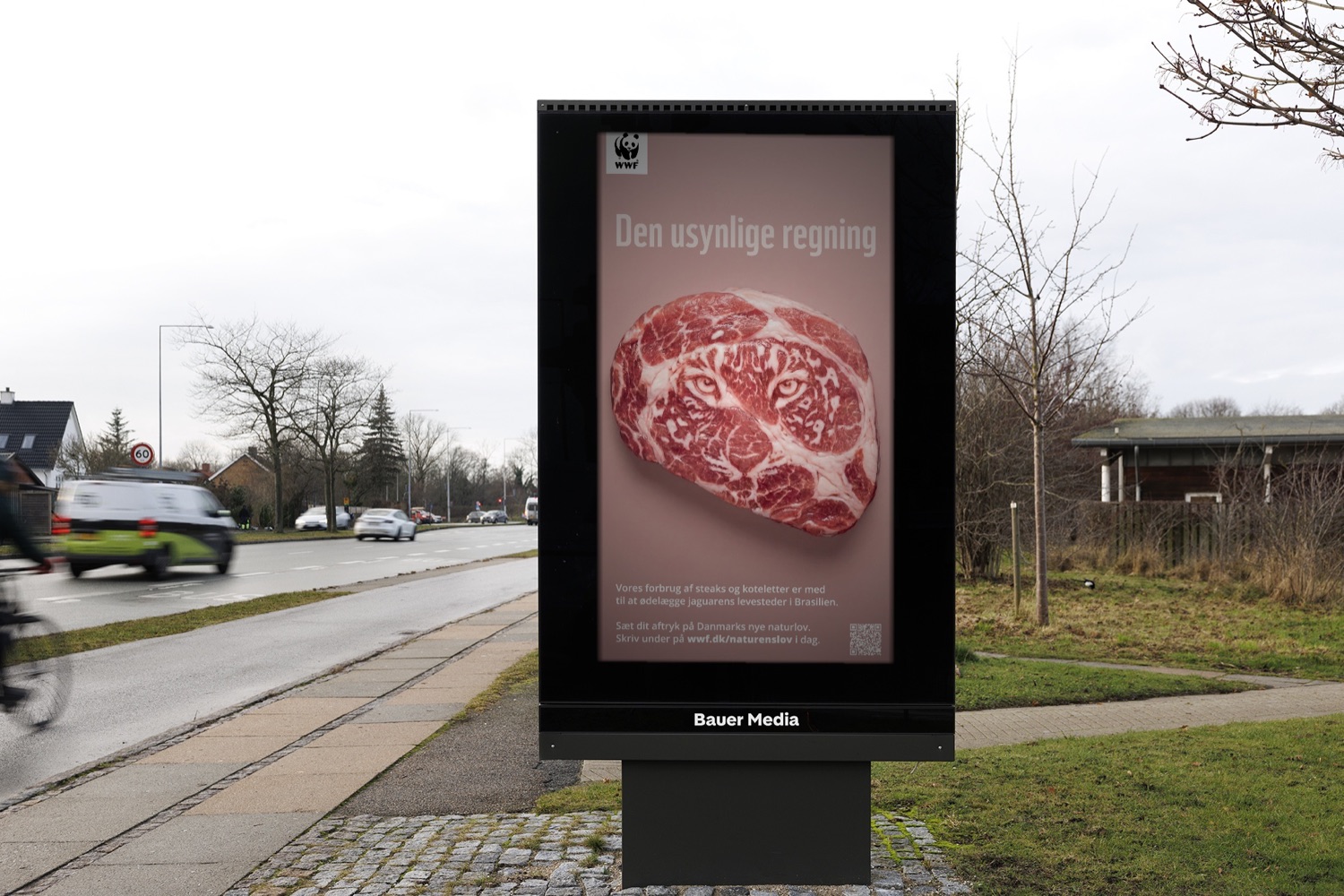 Den Usynlige Regning på en digital billboard langs vejen med forbipasserende biler og en cyklist